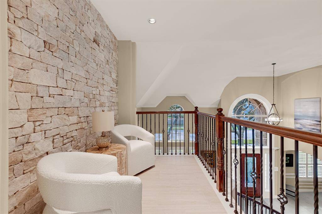 An elegant upper-level interior space with a striking stone accent wall and a sophisticated wrought-iron railing that overlooks a lower level with large arched windows allowing for ample natural light. The space is finished with neutral tones and a classic chandelier.