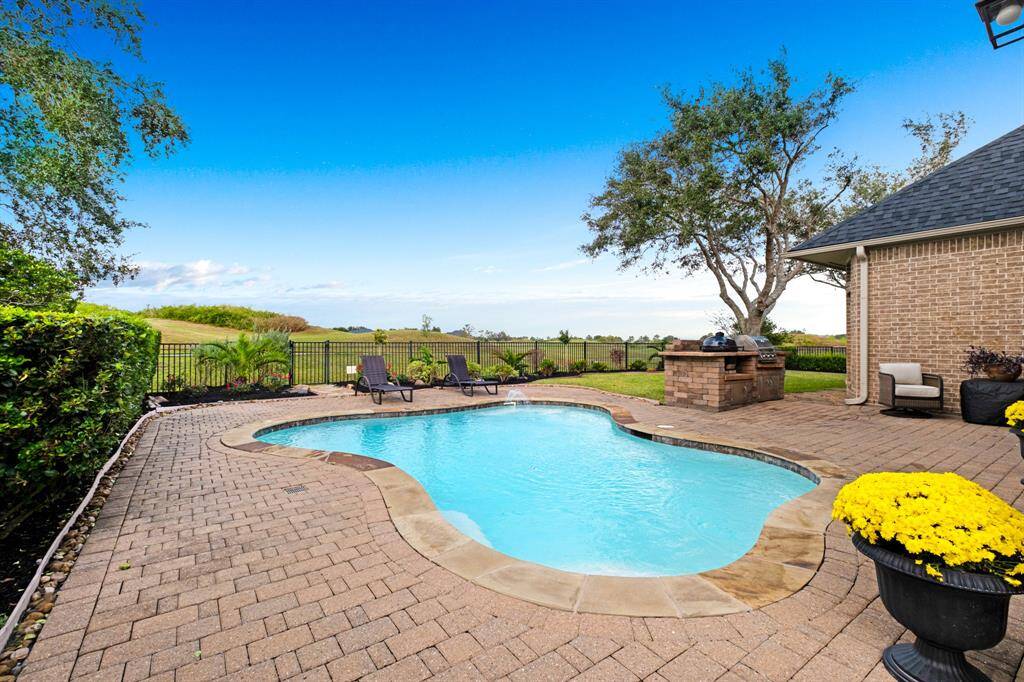 An inviting backyard with a kidney-shaped in-ground pool, a brick paver patio, and a scenic view of an open landscape beyond a metal fence. There is also a covered area with a built-in grill and seating, perfect for outdoor entertaining.