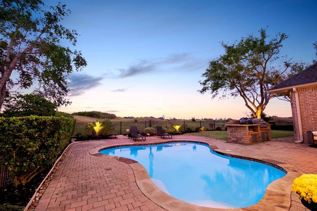 An inviting backyard featuring a curved in-ground pool with a surrounding brick patio, an outdoor grill station, and a serene view of the open sky and landscape beyond the property's wrought-iron fence. Perfect for outdoor entertainment and relaxation.
