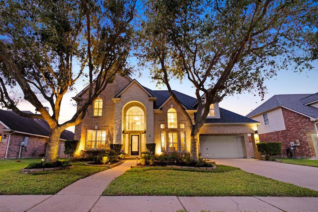 A spacious two-story brick home captured at twilight, featuring an arched entryway, large windows with warm interior lighting, a three-car garage, and mature trees enhancing the curb appeal.