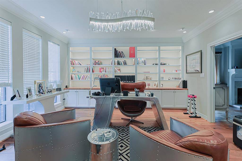 A sleek and modern office space with built-in bookshelves, providing ample storage and display options. Large windows allow for plenty of natural light, while the contemporary lighting fixture adds an elegant touch. The stylish metallic and leather furniture gives the space a sophisticated, industrial-chic vibe.