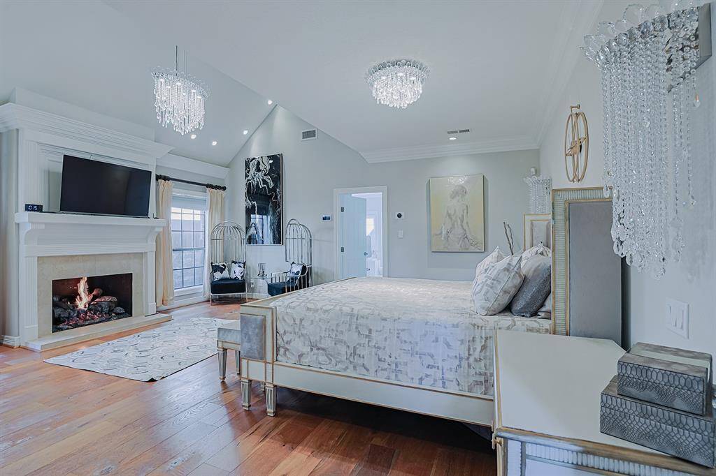 A luxurious and spacious primary suite featuring vaulted ceilings, a cozy fireplace, and a serene seating area. Crystal chandeliers and elegant decor elevate the room’s sophistication, while large windows bring in natural light, creating a warm and inviting retreat.