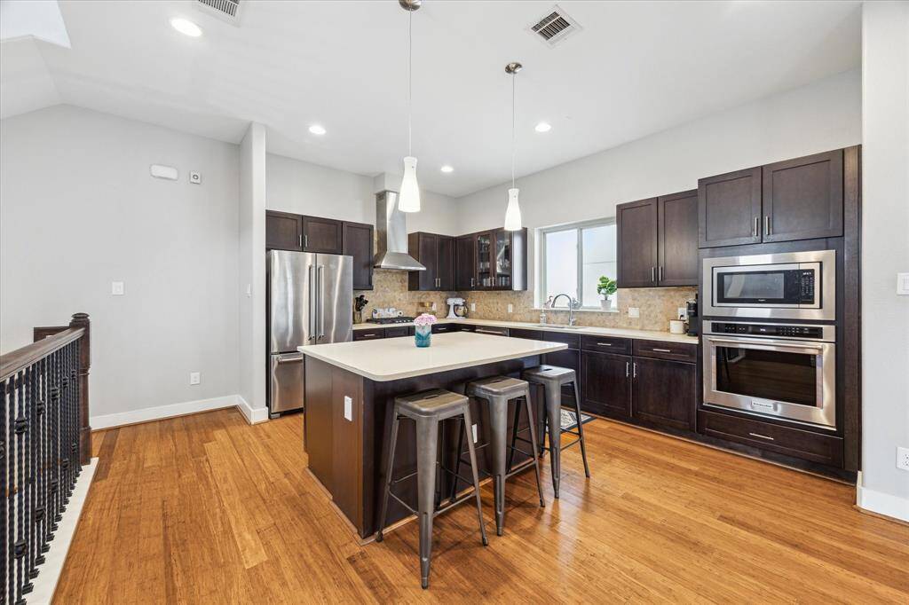 This modern kitchen features sleek dark wood cabinets, stainless steel appliances, and a large island with seating. The open layout is complemented by wood flooring and recessed lighting, creating a bright and inviting space.
