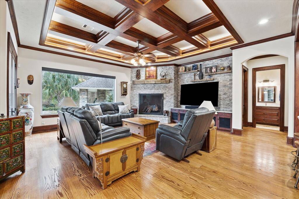 This inviting living room features a coffered ceiling with wood accents and a stone fireplace. It has comfortable seating areas, elegant wood flooring, and a large window offering a view of lush greenery outside, creating a warm and welcoming atmosphere.