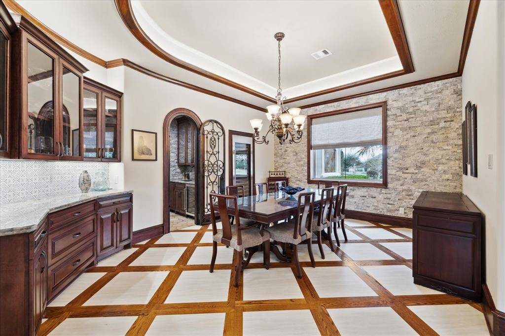 This elegant dining room features rich wooden cabinetry, highlighted by a striking chandelier. It boasts a textured stone accent wall, a large window offering natural light, and intricate ceiling details, creating a warm and inviting atmosphere. It also features a SubZero wine refrigerator