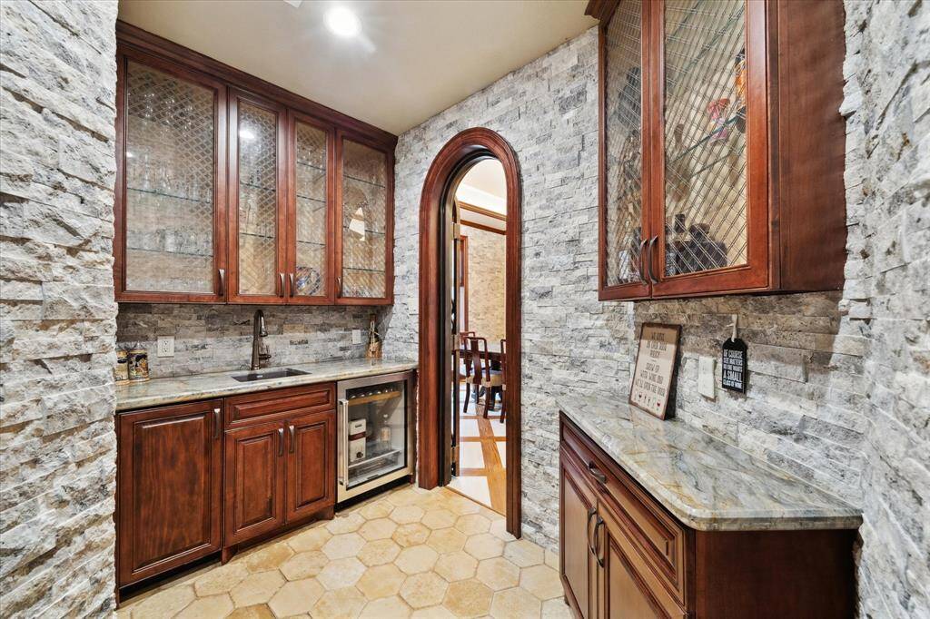 This custom wine grotto with  elegant stone walls, rich wooden cabinetry with glass doors, a small sink, and a built-in mini fridge and ice maker is perfect for entertaining or additional kitchen storage.