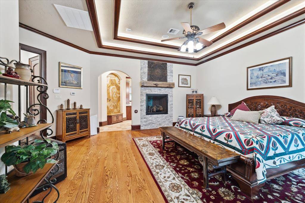 The primary bedroom features hardwood floors, a decorative tray ceiling with a fan, and a cozy fireplace offering a warm and inviting atmosphere.