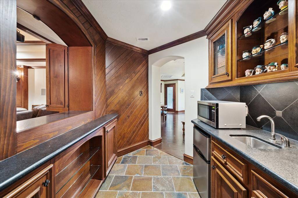 This photo showcases an upstairs wet bar area with rich wood cabinetry, a small sink,a microwave and a passthrough window to the Theater room. The space features tiled flooring and a decorative display shelf, creating a warm and inviting atmosphere perfect for entertaining.