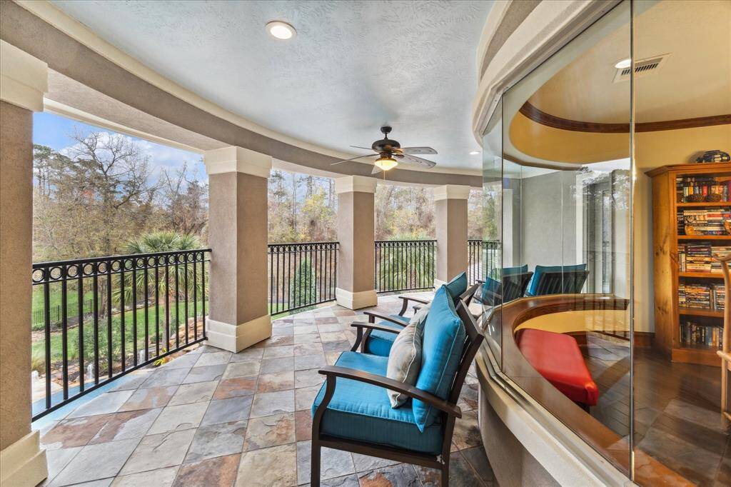 This photo showcases a spacious, covered balcony with elegant tile flooring and comfortable seating area featuring a ceiling fan and a scenic view of lush greenery and the private backyard   The curved glass window offers a glimpse into a cozy indoor space with bookshelves.