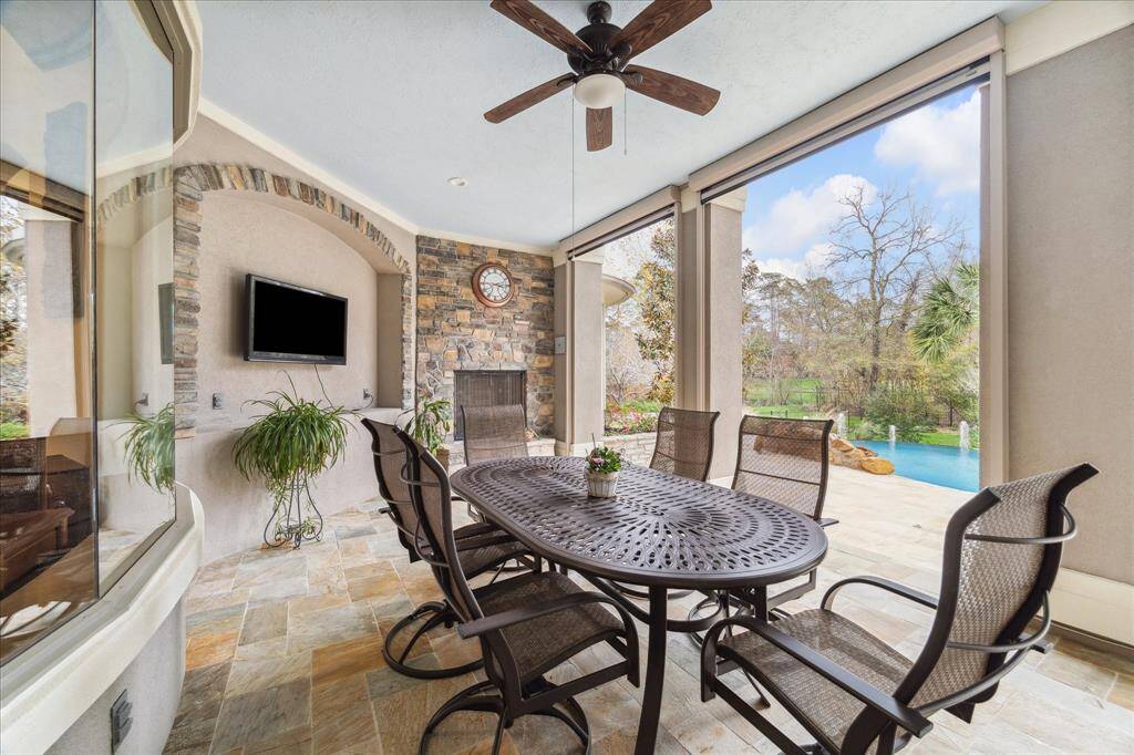 This inviting outdoor patio features a dining area with a ceiling fan, stone fireplace, and mounted TV. There are remote controlled  Solar screens to enclose this back patio. It overlooks the serene pool and lush greenery, perfect for entertaining and relaxation.