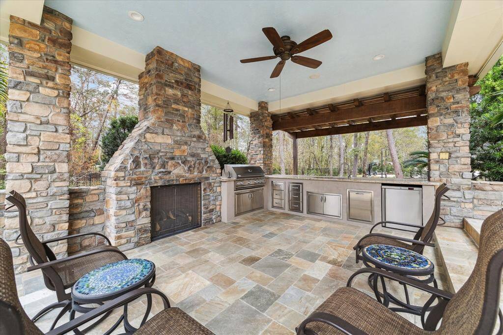 This photo showcases another spacious outdoor patio featuring a stone fireplace, built-in grill, and ample seating. The area has stone pillars, a ceiling fan, and overlooks a wooded area, providing a perfect spot for entertaining or relaxing.