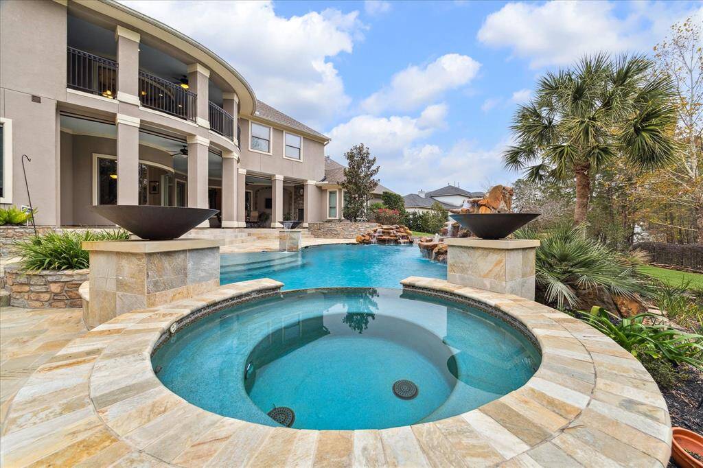 This stunning property features a luxurious outdoor space with a large pool, spa, and elegant stone accents. The home boasts a covered patio and balconies, perfect for relaxation and entertainment, surrounded by lush landscaping and palm trees.