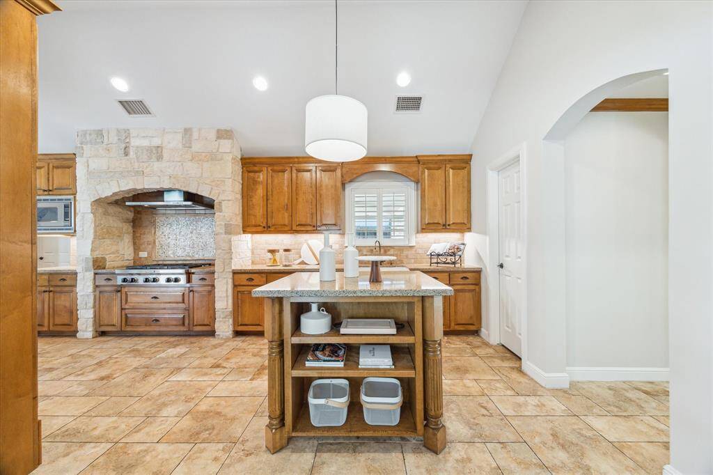 This kitchen features a modern design with wooden cabinetry and stone accents. It has a central island with open shelving, a built-in stove with a stone archway, and ample counter space, creating a warm and inviting atmosphere.
