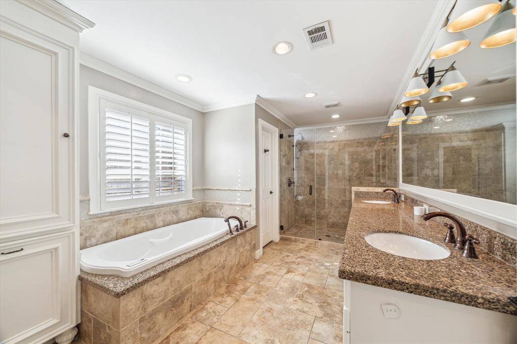 This primary bathroom features a luxurious design with a large soaking bubble jet tub, double vanity with granite countertops, and a spacious glass-enclosed shower. The neutral tile flooring and ample natural light from the window create a bright and inviting atmosphere.