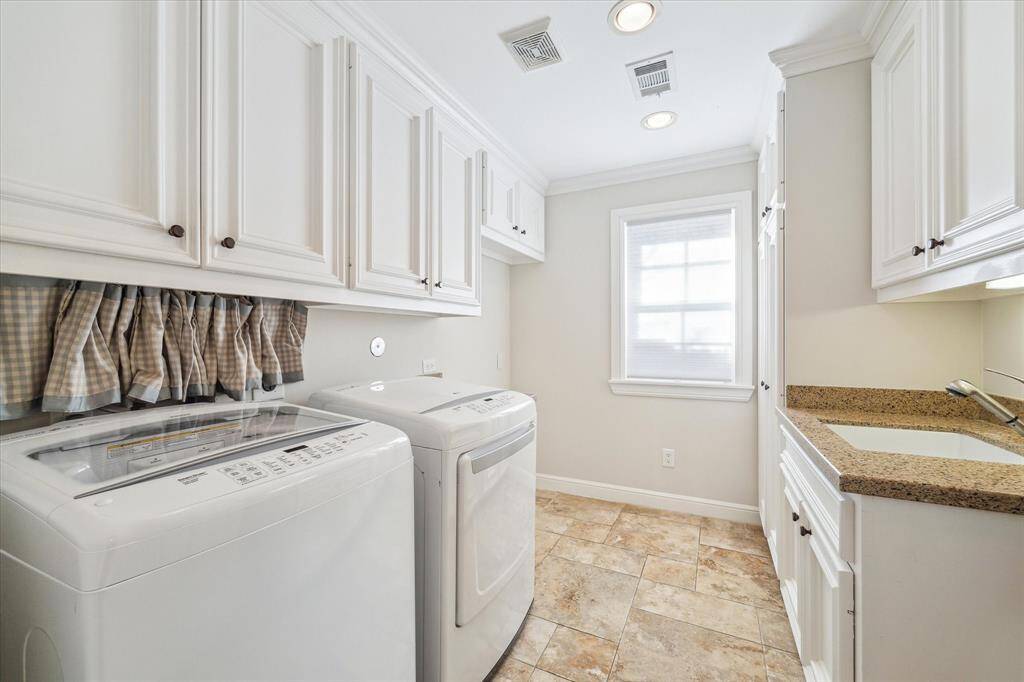 This photo shows a bright, well-organized laundry room featuring a washer and dryer, ample white cabinetry for storage, granite countertop with a sink, and tiled flooring. A window provides natural light.
