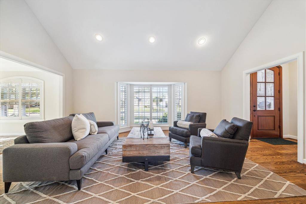This inviting living room features a vaulted ceiling with recessed lighting, a large window with plantation shutters, and warm hardwood floors.