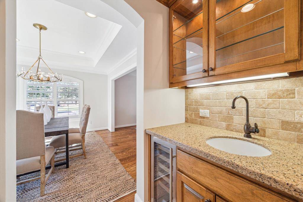 This photo showcases a bright dining area with a large windows, elegant chandelier, and hardwood floors. Adjacent is a cozy wet bar featuring granite countertops, a sink, and glass cabinetry and a wine chiller —ideal for entertaining.