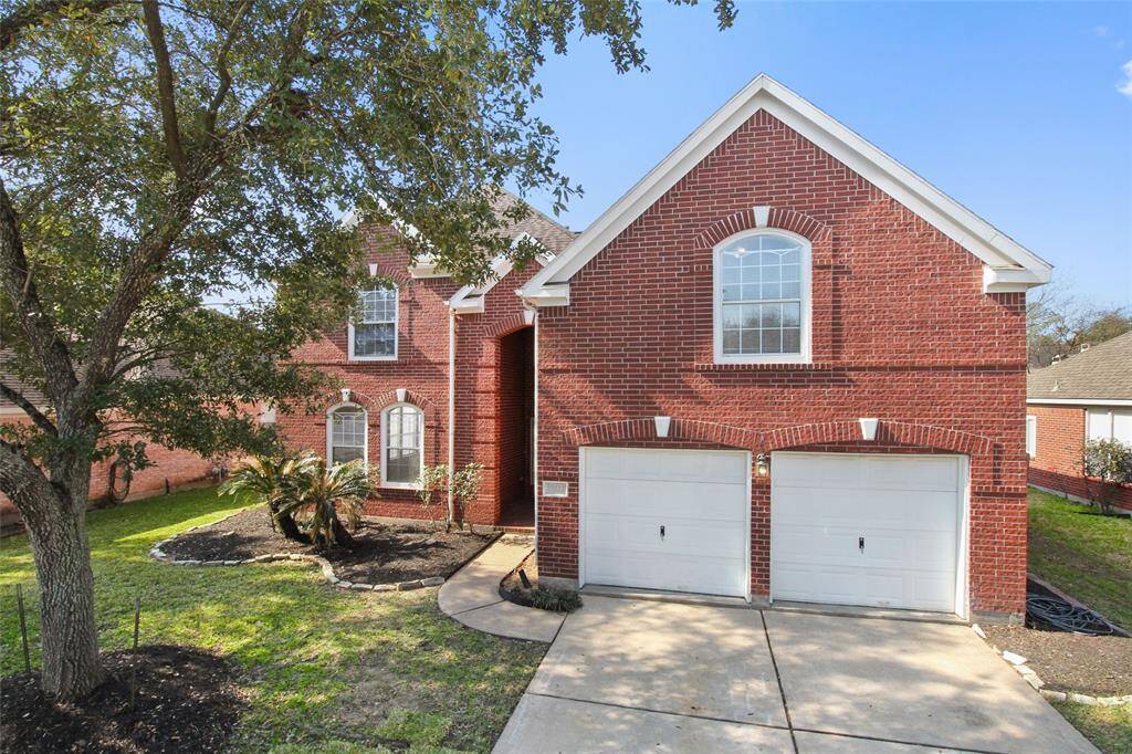 This is a charming red-brick, two-story home with a double garage. It features a well-maintained front yard with mature trees and landscaping. The house has large, arched windows and a welcoming entrance.