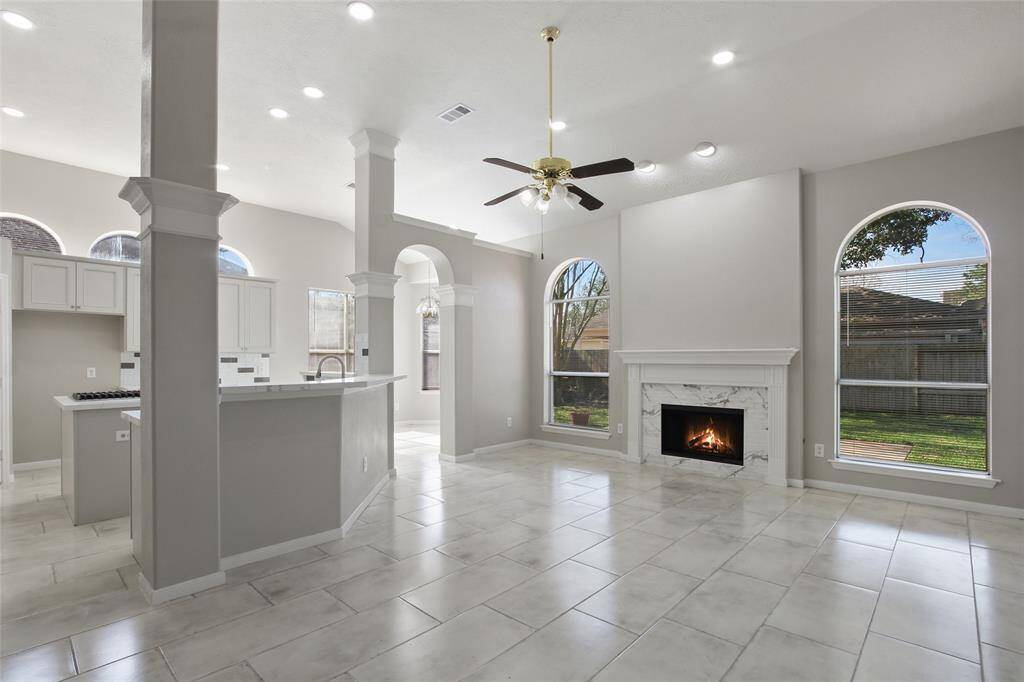 This spacious living area features high ceilings, large arched windows for natural light, and a cozy fireplace. The open layout seamlessly connects to the modern kitchen, which includes elegant cabinetry and a breakfast bar. The neutral palette and tiled flooring enhance the room's brightness and elegance.