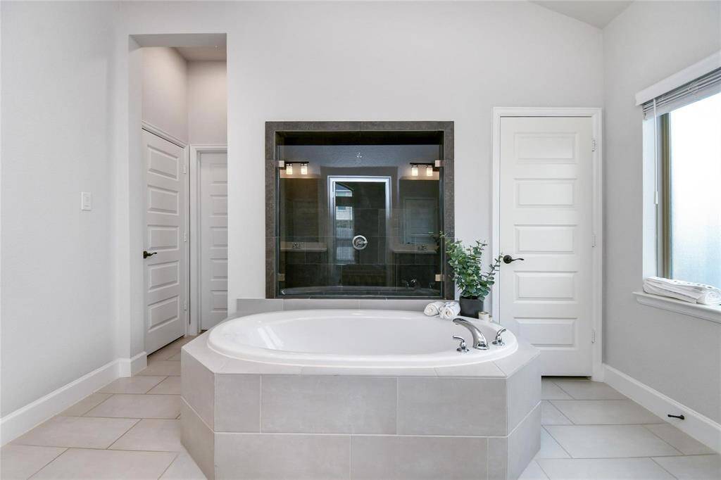 This bathroom features a spacious soaking tub with modern fixtures, a separate glass-enclosed shower, light tile flooring, and ample natural light from a large window.