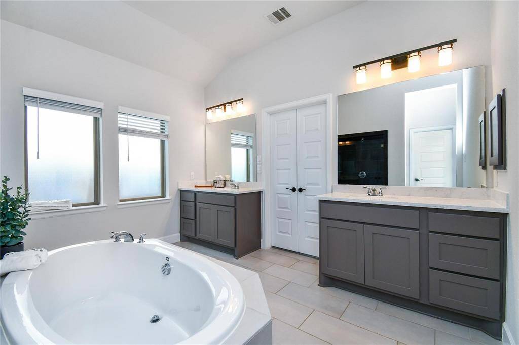 This bathroom features a spacious layout with a large soaking tub, dual vanities with modern fixtures, and ample natural light from two frosted windows. The sleek cabinetry and neutral tiles create a clean, contemporary look.