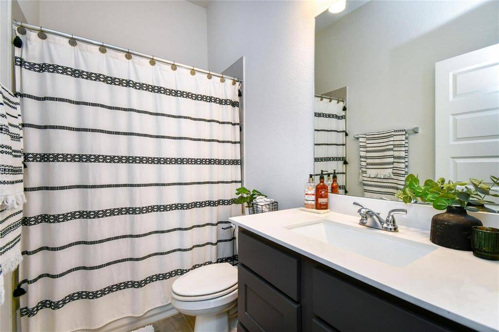 This bathroom features a modern design with a sleek vanity and sink, complemented by a large mirror. The shower curtain has a stylish black and white pattern, and there are decorative plants adding a touch of greenery.