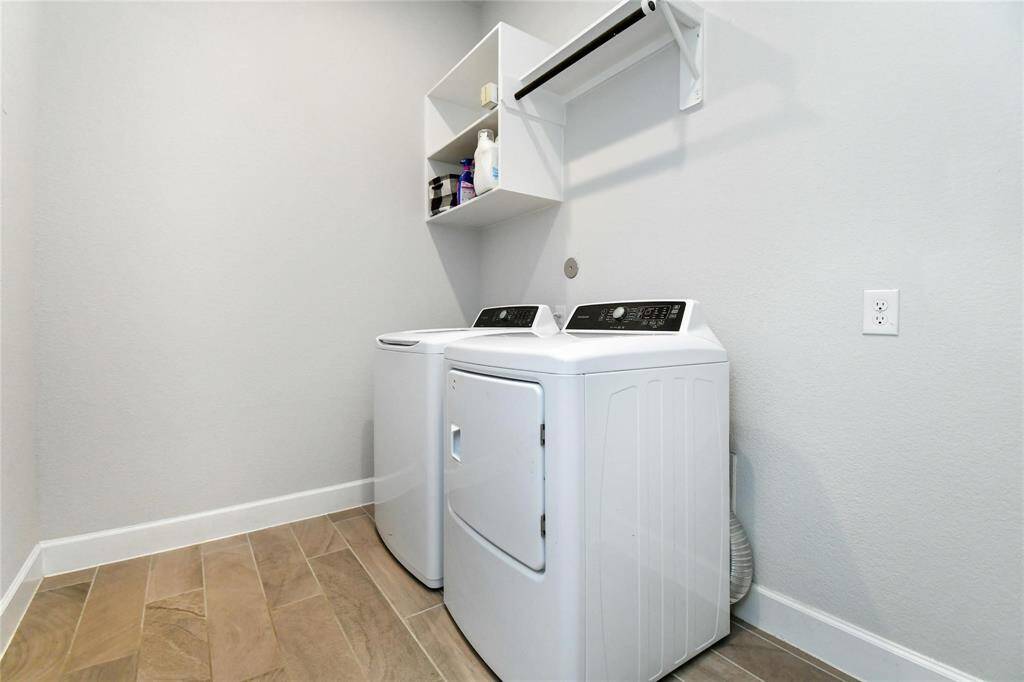 This photo shows a modern laundry room with a washer and dryer set, light-colored walls, and wood-like tile flooring. There's overhead shelving for storage and a wall-mounted drying rod.