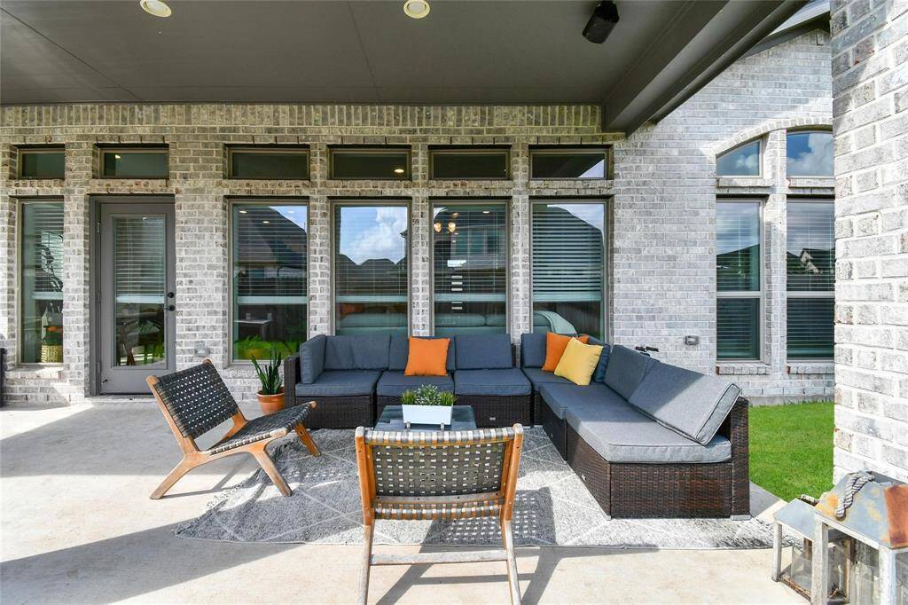 This photo showcases a stylish outdoor patio featuring a large, comfortable sectional sofa with colorful cushions, two modern chairs, and a small table on a patterned rug. The brick exterior and large windows create an inviting atmosphere, perfect for relaxation and entertaining.