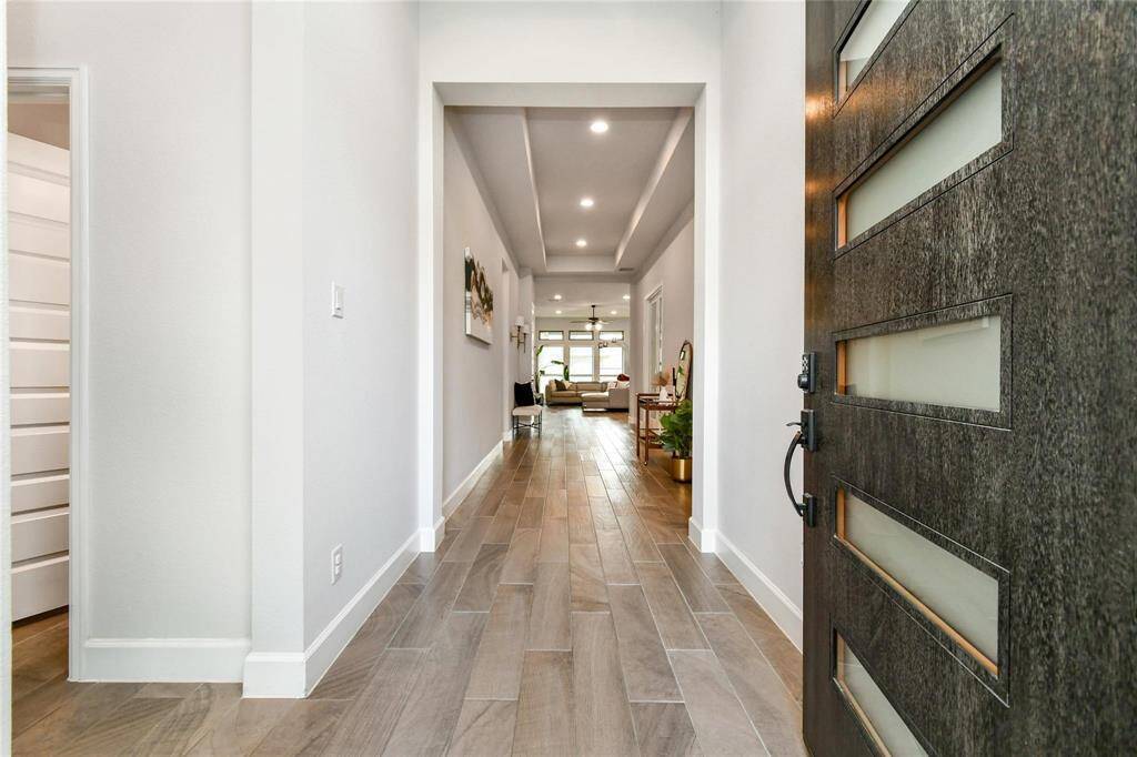 This photo showcases a modern entryway with a sleek, dark front door featuring frosted glass panels. The hallway has light wood-look tile flooring, leading to a bright, inviting living area. The space is well-lit with recessed lighting and has a clean, contemporary design.