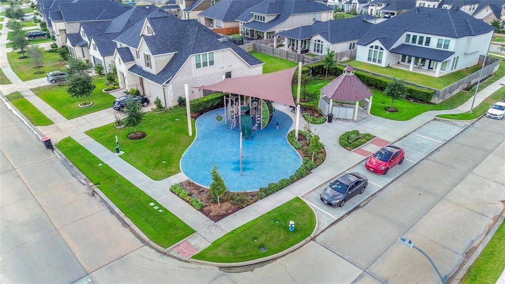 This aerial photo showcases a modern, family-friendly neighborhood with spacious homes and well-maintained lawns. A central playground with a canopy and parking area highlights the community's amenities, surrounded by attractive residential properties.