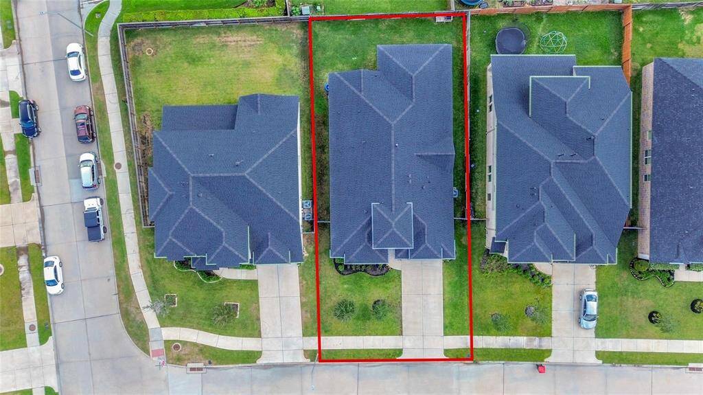 This aerial view shows a modern two-story home with a dark roof, highlighted within a red boundary. It features a driveway and a neatly maintained lawn both in front and back. The property is situated in a suburban neighborhood with adjacent homes and a clean streetscape.