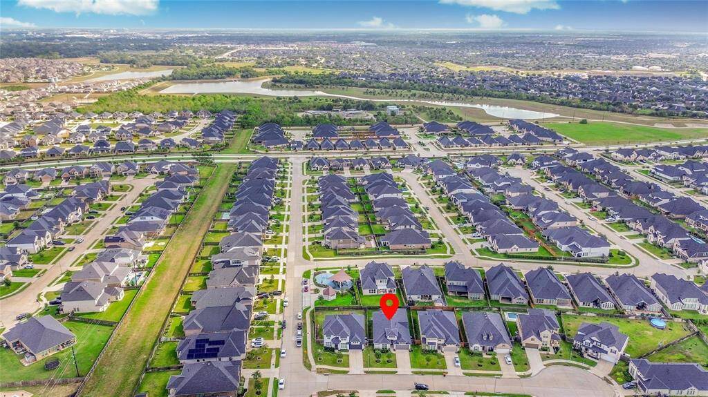 This aerial photo shows a large residential neighborhood with rows of similar, well-maintained homes. Streets are neatly arranged, and the area is surrounded by greenery and open space, creating a suburban feel. A red marker highlights a specific property, indicating its location within the community.