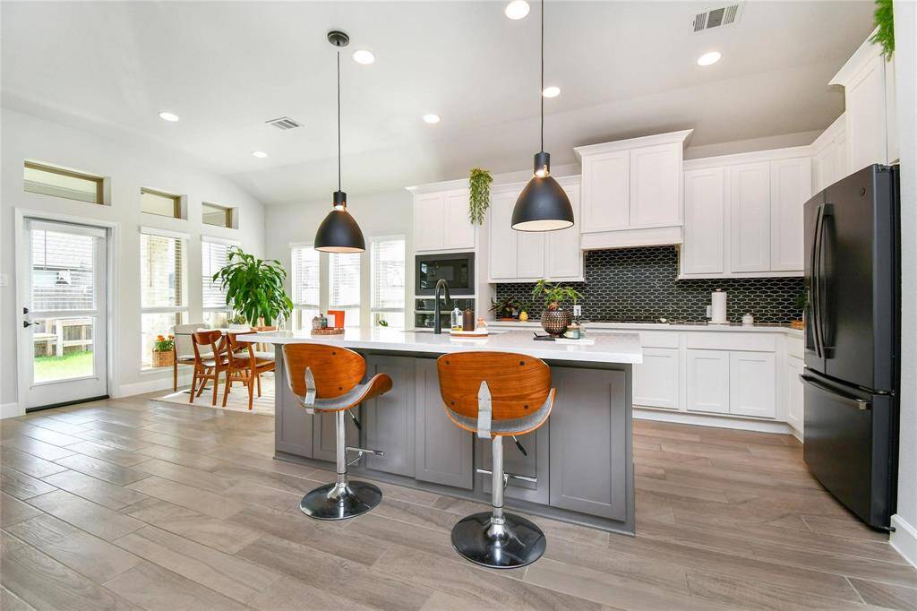 This modern kitchen features sleek white cabinetry, a stylish black backsplash, and a central island with seating. It includes stainless steel appliances, pendant lighting, and ample natural light from large windows. The open layout provides a spacious dining area and access to the backyard.