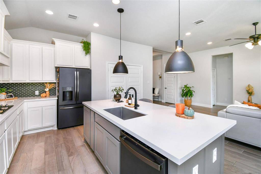 This modern kitchen features a spacious island with a sink, sleek black appliances, and white cabinetry. Pendant lighting and a stylish backsplash add a contemporary touch, while the open layout connects to a cozy living area with neutral tones and wood flooring.