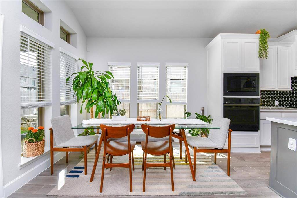 Bright dining area with a glass table and modern chairs, surrounded by large windows. The space features a mix of natural light, greenery, and a sleek kitchen with white cabinets and built-in appliances, creating a fresh and inviting atmosphere.
