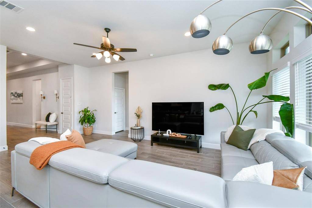 This spacious living room features a modern design with a large sectional sofa, sleek ceiling fan, and stylish floor lamp. The room is bright with natural light from multiple windows, and it includes a large TV and tasteful decor with plants and minimalist accents.