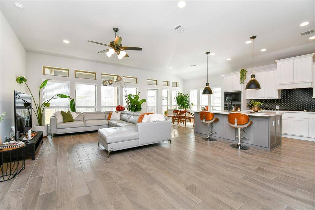 This photo showcases a spacious, open-concept living area with modern design. It features a large kitchen island with seating, sleek white cabinetry, and pendant lighting. The living room has ample natural light from multiple windows, a ceiling fan, and contemporary furnishings. The space is enhanced by wood-look flooring and greenery, creating a welcoming and stylish atmosphere.