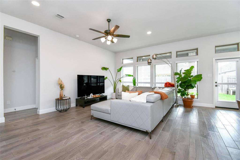 This bright and spacious living room features modern wood flooring, neutral walls, and large windows for plenty of natural light. It includes a stylish ceiling fan, a sleek sectional sofa, and a TV setup, creating a comfortable and inviting space. The room also has access to the backyard through a glass door.