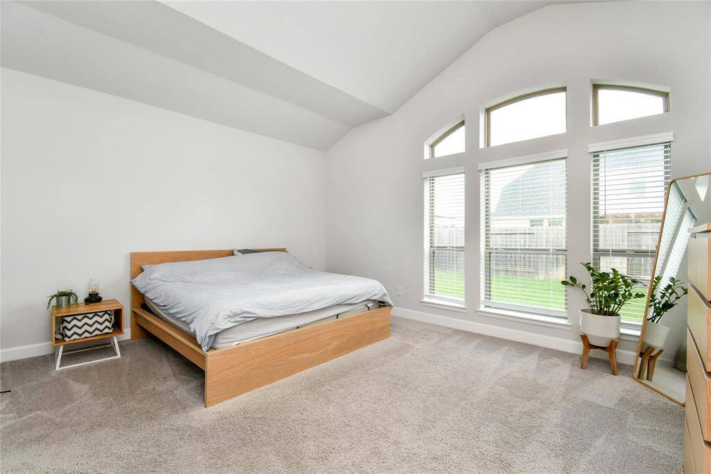 This spacious bedroom features a high ceiling and large arched windows, providing ample natural light. It includes a modern wooden bed, cozy carpeting, and a touch of greenery, creating a serene and inviting atmosphere.