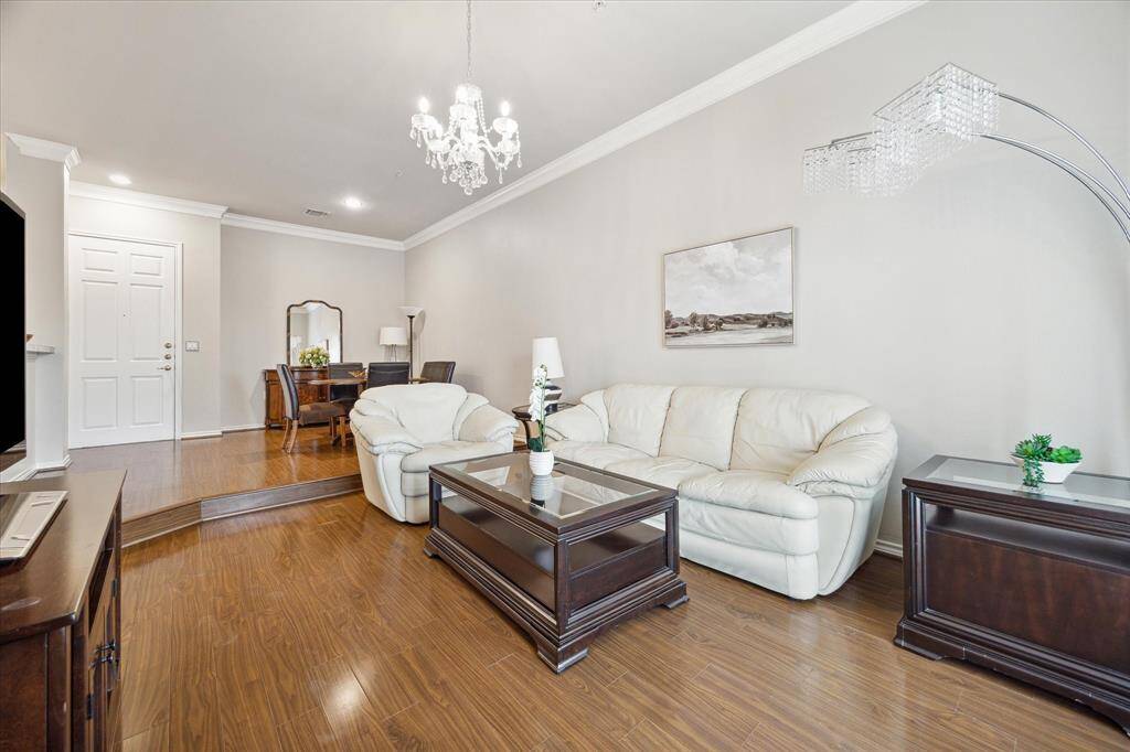 This inviting living room features hardwood floors, a cozy white sofa set, and elegant dark wood furniture. A chandelier adds a touch of luxury, while the open layout leads to a dining area, creating a spacious and welcoming atmosphere.