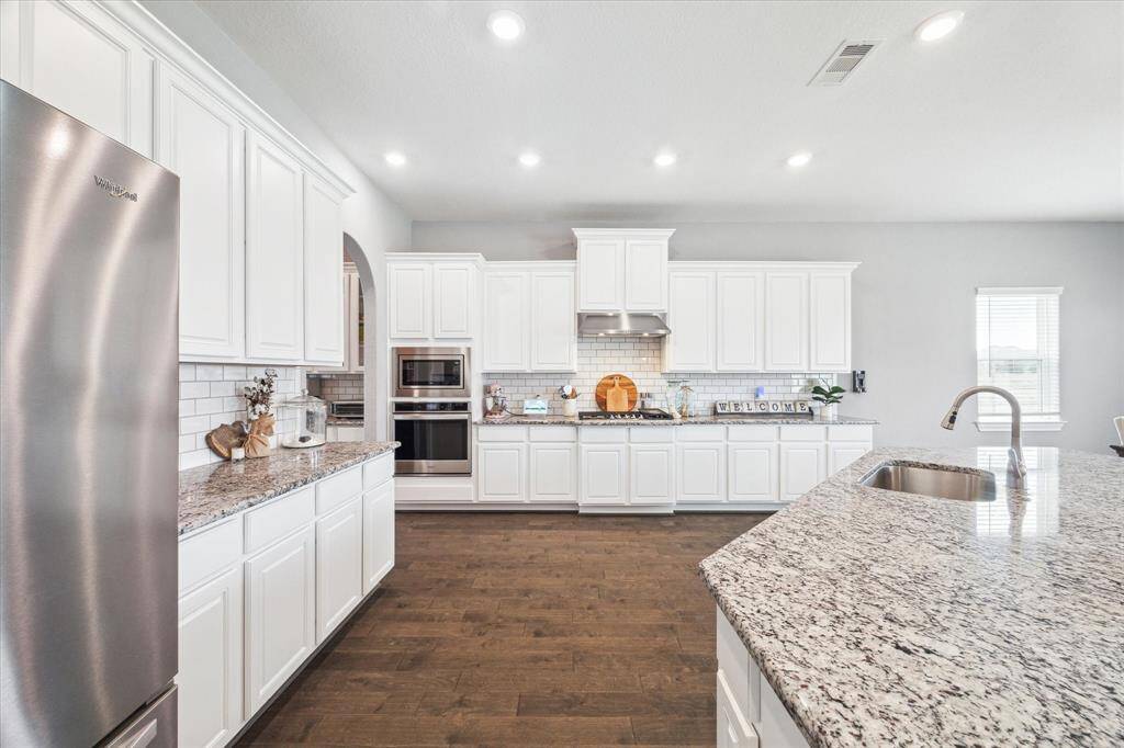 Chef's kitchen features modern stainless steel appliances, granite countertops, and ample white cabinetry. The open layout is enhanced by a large island with a sink. Warm wood flooring and subway tile backsplash add a stylish touch.