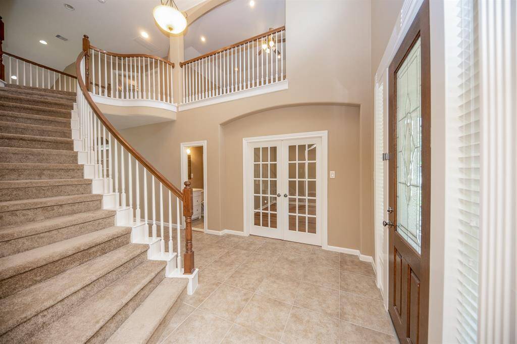 This photo showcases a spacious, bright entryway featuring a grand staircase with carpeted steps and a wooden handrail. The tiled floor leads to elegant French doors, and natural light enhances the warm, neutral tones of the walls.