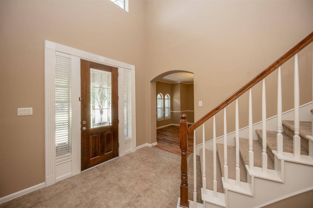 The entryway features a tiled floor, a wooden front door with decorative glass, and a staircase with carpeted steps and wooden railing. There's an arched opening leading to the dining room with elegant flooring and large windows.