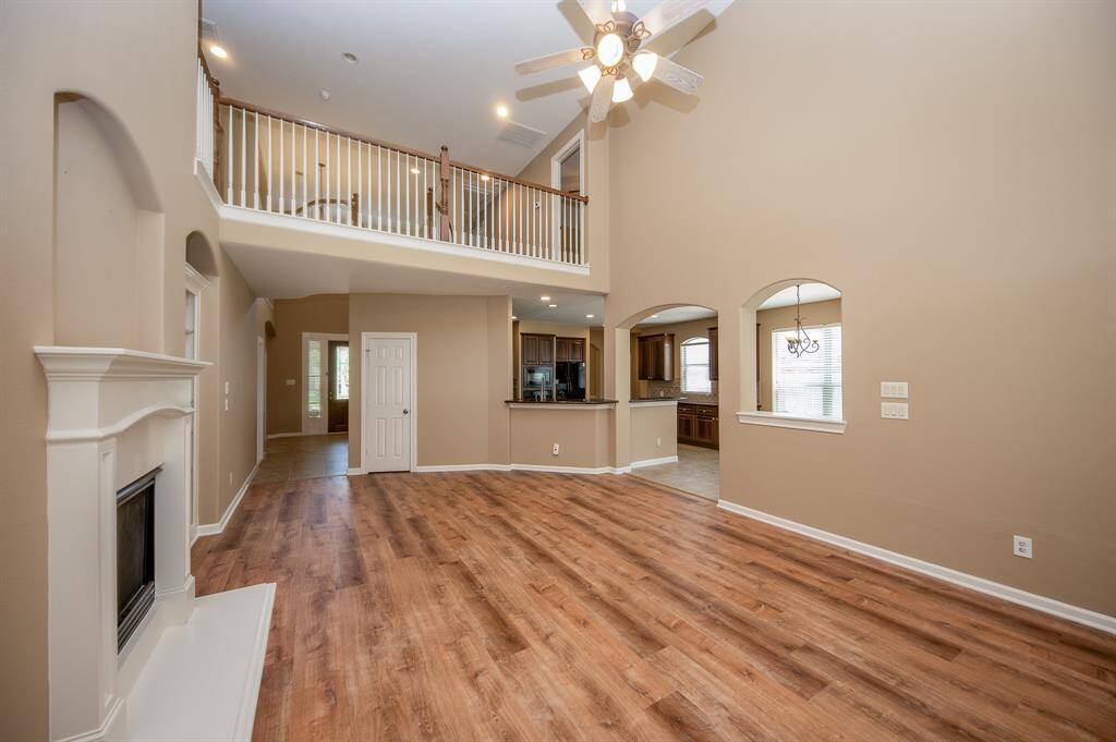 This spacious living area features a high ceiling with a fan, a cozy fireplace, and beautiful flooring. It opens to an upstairs landing and connects to a kitchen and dining area, creating a welcoming and open atmosphere.