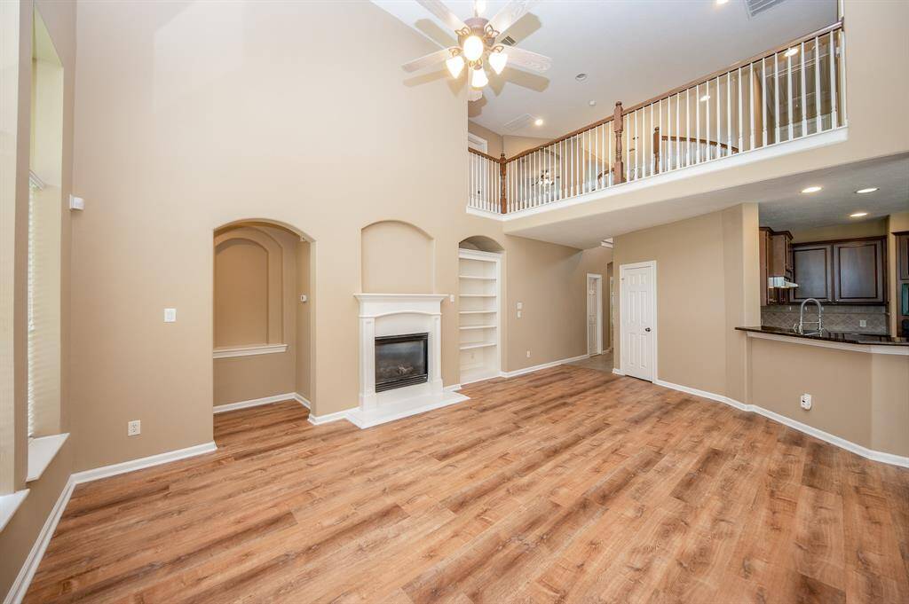 This spacious living area features high ceilings, gorgeous flooring, and a cozy fireplace. It includes built-in shelving, a ceiling fan, and an open layout leading to a modern kitchen with a breakfast bar. The upper-level balcony adds an elegant touch.