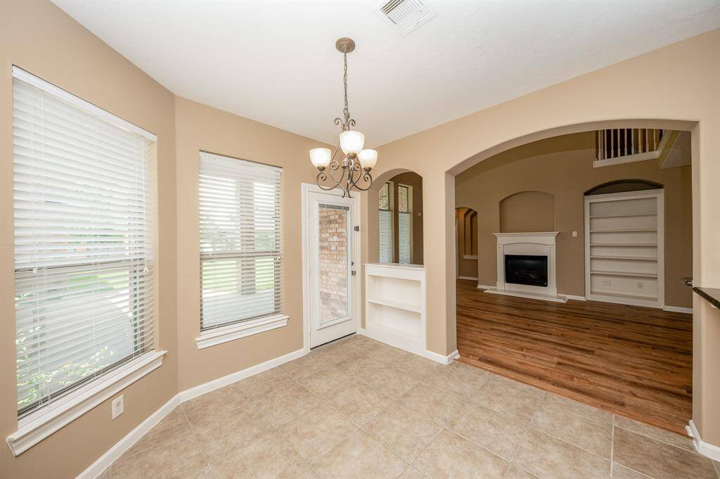 This photo features a bright dining area with large windows and tile flooring, leading to a cozy living room with complimenting flooring, built-in shelves, and a fireplace. An elegant chandelier adds charm, and an archway connects the spaces.