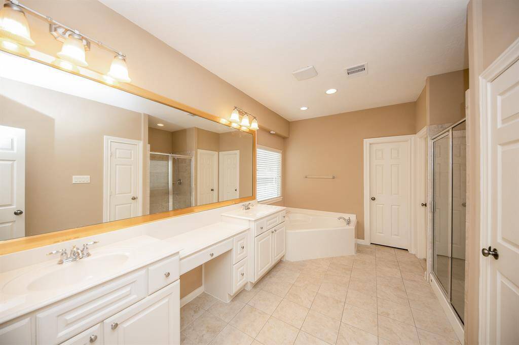 This spacious bathroom features dual sinks with ample counter space, a large mirror, and elegant lighting. It includes a separate shower, a cozy corner bathtub, and neutral tile flooring, offering a clean and bright atmosphere.