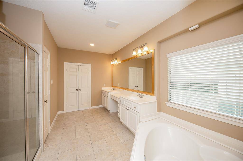 This spacious bathroom features a large vanity with dual sinks, a walk-in shower, and a separate bathtub. It has ample natural light from a window with blinds, tiled flooring, and a neutral color palette.