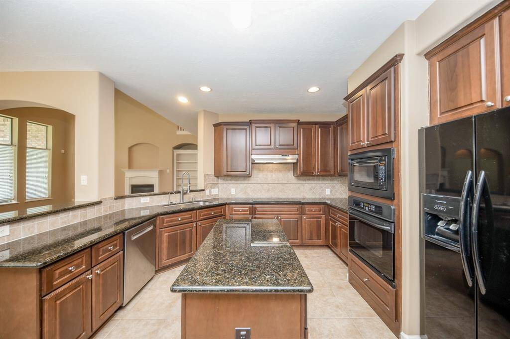 Spacious kitchen with dark granite countertops, rich wooden cabinets, and a central island. It features modern appliances, including a double oven and a large refrigerator, with ample natural light from adjacent windows.