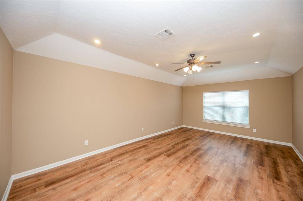 This spacious bedroom features warm beige walls and elegant flooring, complemented by recessed lighting and a ceiling fan. A large window allows for ample natural light.